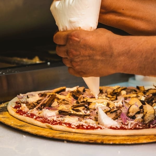 Hands squeezing a piping bag of cheese onto a pizza with mushrooms and red onions.
