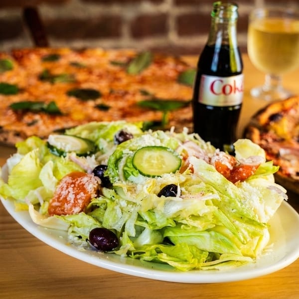 Fresh salad with two pizzas, a Coke bottle, and white wine on a wooden table.