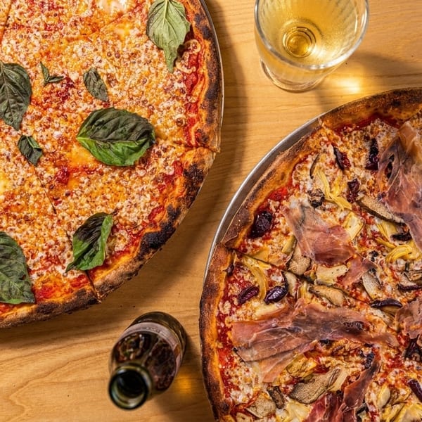Two pizzas, one Margherita and one with prosciutto, artichokes, and olives, on a wooden table.