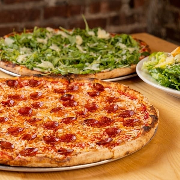 Two pizzas, one pepperoni and one with arugula, alongside a Caesar salad on a wooden table.